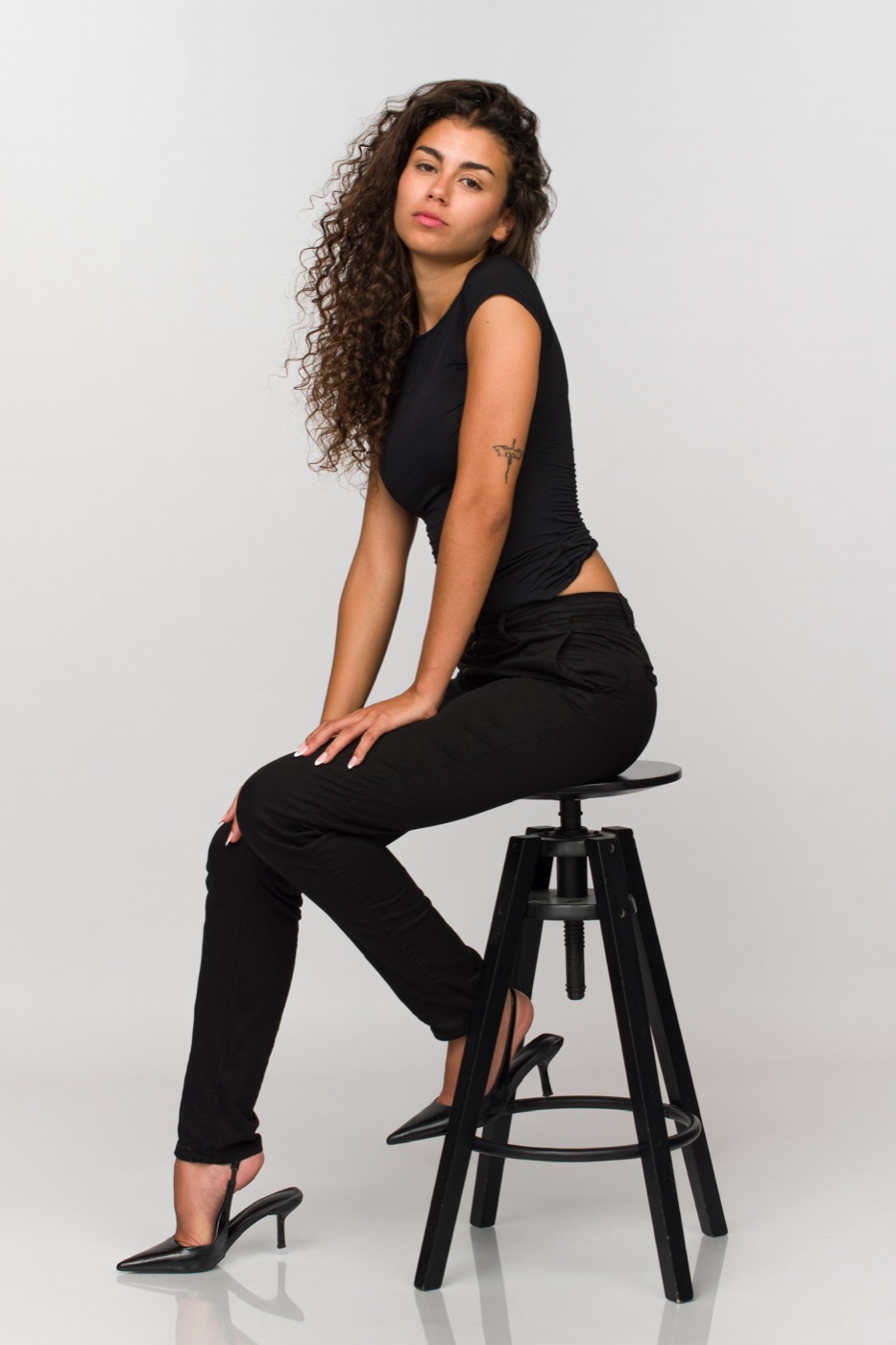 Vitoria sentada en un taburete, camiseta y pantalón negros, tacón